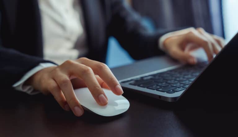 A close-up of a person's hands using a white computer mouse and a laptop to write blog headlines for beginners.